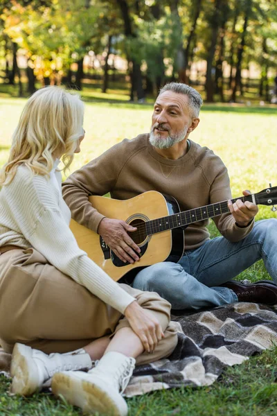 Feliz hombre de mediana edad con barba gris tocando la guitarra acústica cerca de la esposa rubia en el parque - foto de stock