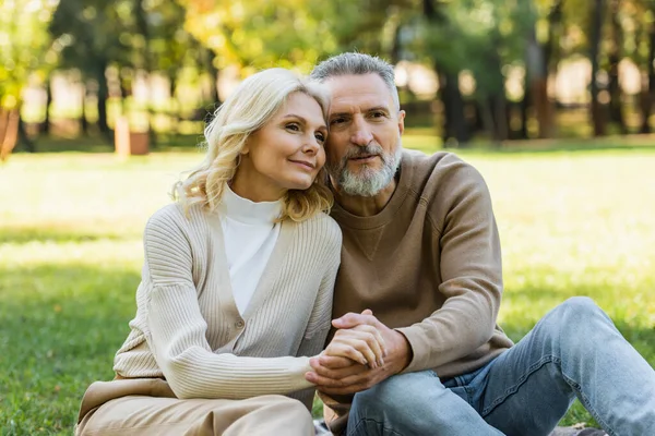 Portrait of charming middle aged couple holding hands while sitting together in green park during springtime — Stock Photo