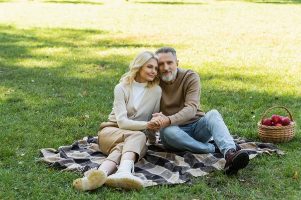 Charmantes Paar mittleren Alters hält Händchen, während es im Frühling auf einer Decke im grünen Park sitzt — Stockfoto