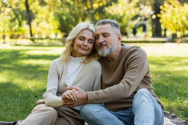 Pleased middle aged couple sitting together with closed eyes in green park during springtime — Stock Photo