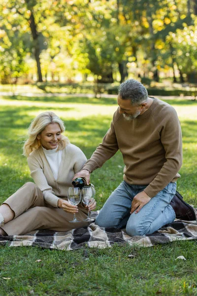 Middle aged man pouring wine into glass near happy wife during picnic in park — Stock Photo