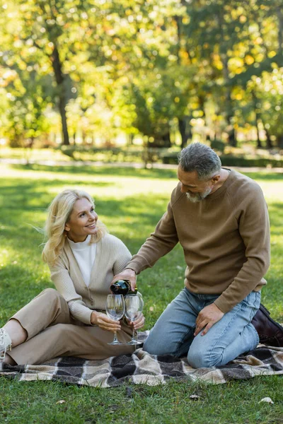 Middle aged man pouring wine into glass near joyful wife during picnic in park — Stock Photo