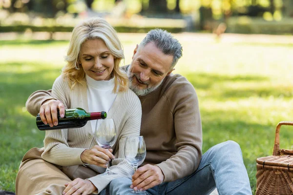 Homem de meia idade feliz derramando vinho em vidro perto da esposa alegre durante o piquenique no parque — Fotografia de Stock