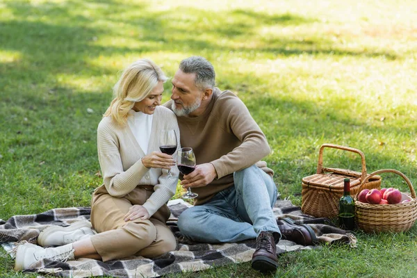 Barbudo hombre de mediana edad tintineo vasos de vino con rubia y feliz esposa durante el picnic - foto de stock