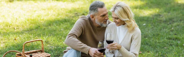 Barbudo hombre de mediana edad tintineo vasos de vino con rubia y alegre esposa durante el picnic, pancarta - foto de stock
