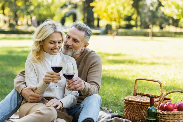 Pleased middle aged man clinking glasses of wine with blonde and happy wife during picnic — Stock Photo