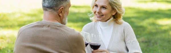 Pleased middle aged couple clinking glasses with red wine in green park, banner — Stock Photo