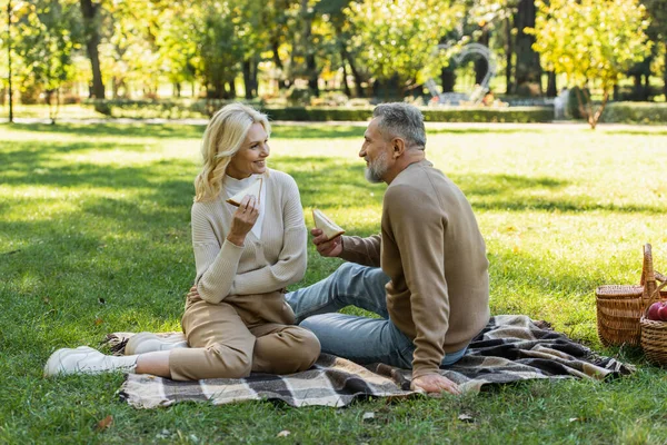 Happy middle aged couple holding tasty sandwiches during picnic in green park — Stock Photo
