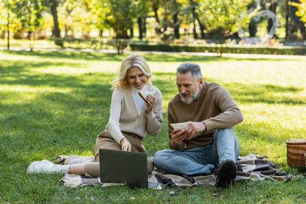 Heureux couple d'âge moyen manger de savoureux sandwichs club et regarder un film sur ordinateur portable pendant le pique-nique dans le parc vert — Photo de stock