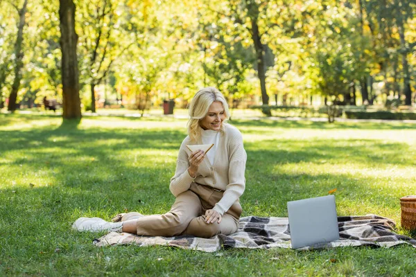 Cheerful middle aged woman with blonde hair eating sandwich and watching movie on laptop during picnic in park — Stock Photo