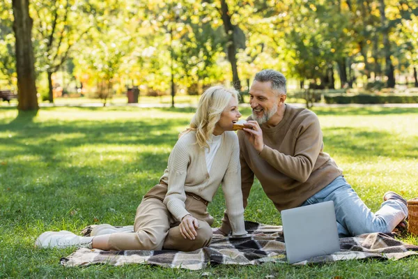 Happy middle aged man feeding blonde wife with tasty sandwich during picnic in park — Stock Photo