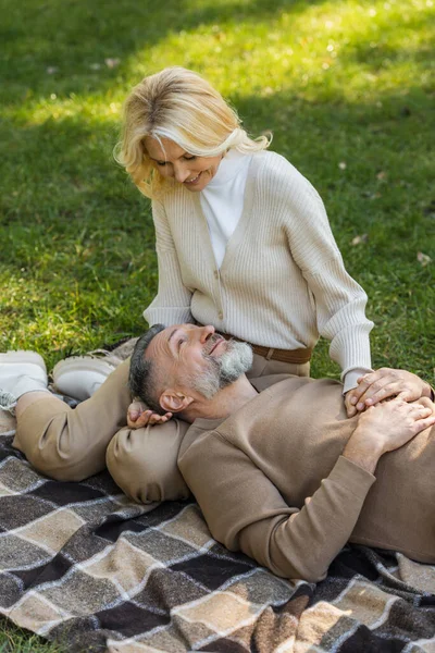 Pleased middle aged man lying on laps of blonde and happy wife during picnic in park — Stock Photo