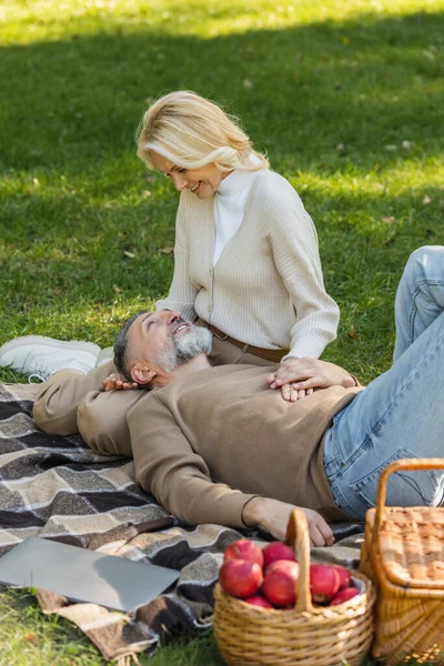 Pleased middle aged man lying on laps of blonde and cheerful wife during picnic in park — Stock Photo