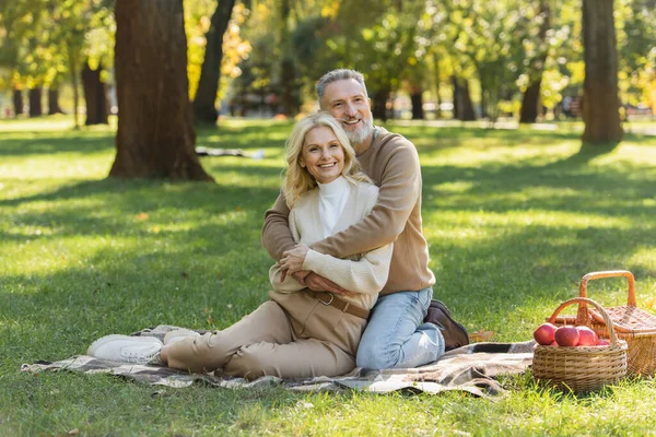 Glücklicher Mann mittleren Alters mit grauem Bart umarmt charmante blonde Frau beim Picknick im Park — Stockfoto