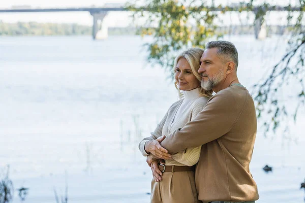 Весёлый мужчина средних лет с седой бородой обнимает счастливую блондинку жену возле реки в парке — стоковое фото