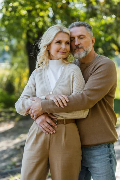 Cheerful middle aged man with closed eyes hugging happy wife in park — Stock Photo