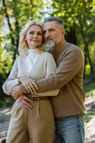 Бородатий чоловік середнього віку обіймає чарівну блондинку дружину в парку — стокове фото