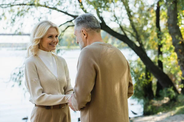 Счастливая женщина средних лет, держась за руки с мужем, глядя друг на друга возле озера в парке — стоковое фото