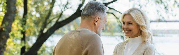 Happy middle aged woman looking at bearded husband near lake in park, banner — Stock Photo