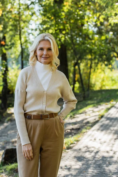 Happy middle aged woman with blonde hair posing with hand on hip in green park — Stock Photo