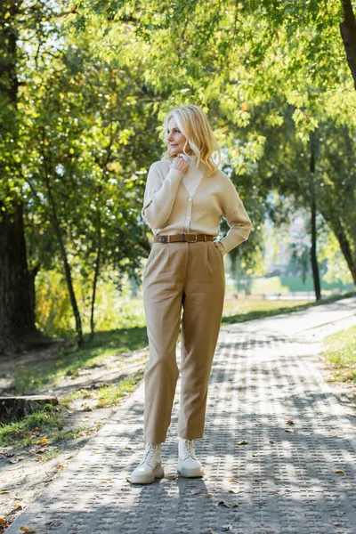 Full length of cheerful middle aged woman with blonde hair posing with hand on hip in green park — Stock Photo