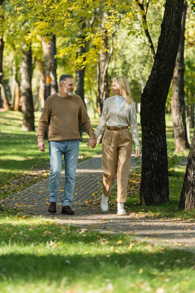 Full length of stylish middle aged couple holding hands and smiling while walking together in park during springtime — Stock Photo