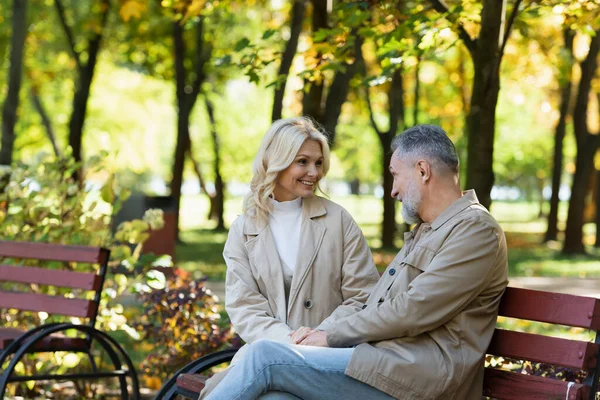 Alegre pareja de mediana edad hablando mientras está sentado en el banco en verde parque - foto de stock