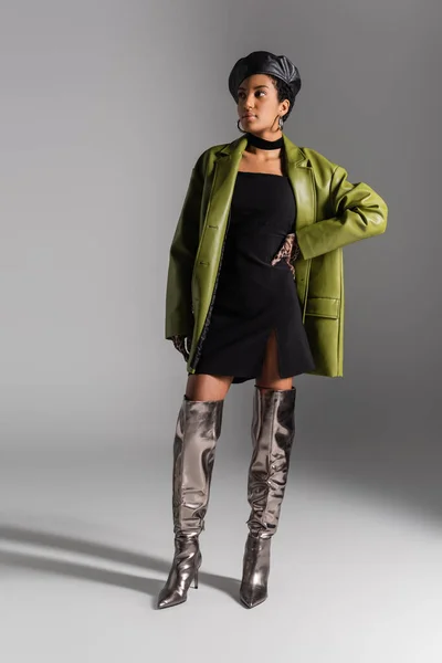 Donna afroamericana alla moda in stivali lucidi e cappotto di pelle in posa su sfondo grigio — Foto stock