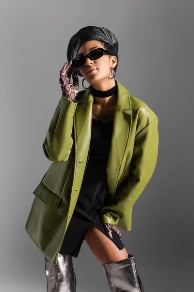 Modèle afro-américain à la mode en manteau de cuir touchant les lunettes de soleil isolées sur gris — Photo de stock