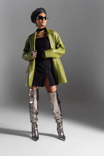 Longitud completa del modelo afroamericano de moda en abrigo de cuero y gafas de sol sobre fondo gris — Stock Photo