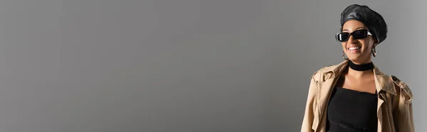 Femme afro-américaine insouciante et à la mode en lunettes de soleil et trench coat souriant isolé sur gris, bannière — Photo de stock