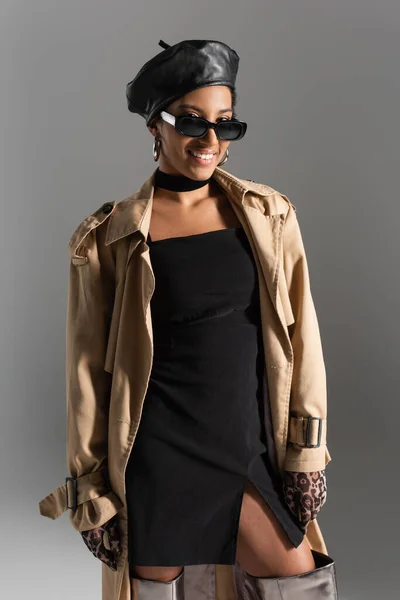 Sorrindo mulher americana africana em óculos de sol e casaco de trincheira olhando para a câmera isolada em cinza — Fotografia de Stock