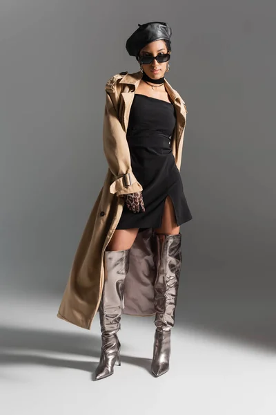 Повна довжина стильної афро-американської жінки в блискучі чоботи і траншеї пальто стоячи на сірому фоні — стокове фото