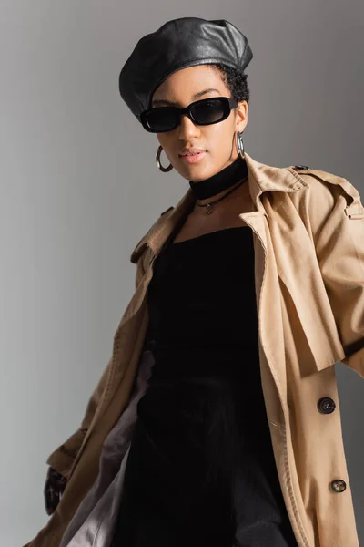 Trendy african american model in sunglasses and trench coat standing isolated on grey — Stock Photo