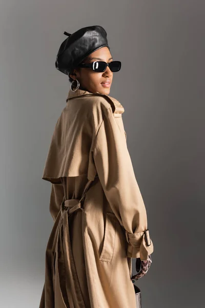 Modelo americano africano na moda em boina e casaco de trincheira de pé isolado em cinza — Fotografia de Stock