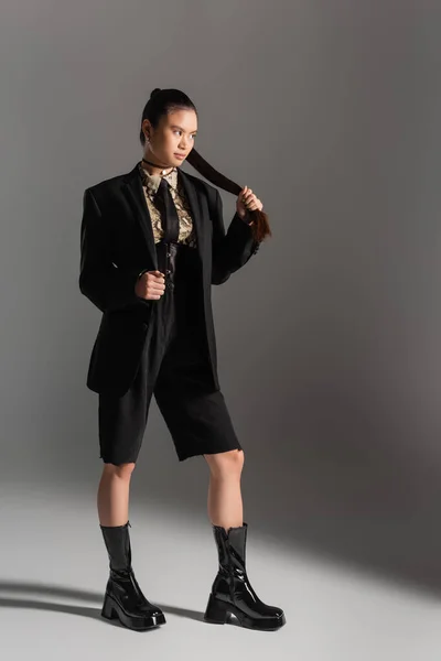 Longitud completa de mujer asiática con estilo en chaqueta y pantalones cortos tocando el pelo sobre fondo gris - foto de stock