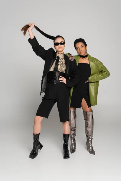 Full length of stylish interracial girlfriends in jackets and boots posing on grey background — Stock Photo
