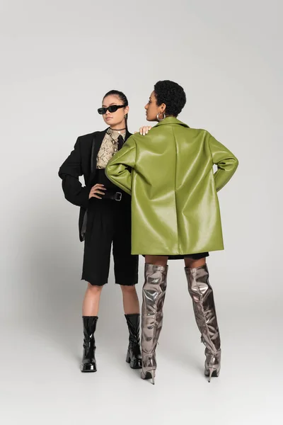 Full length of stylish asian model posing near african american friend in leather coat on grey background — Stock Photo