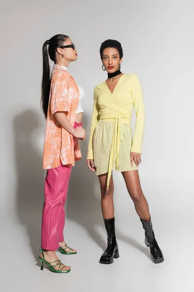 Comprimento total de modelos inter-raciais elegantes em roupas de primavera brilhante posando em fundo cinza — Fotografia de Stock