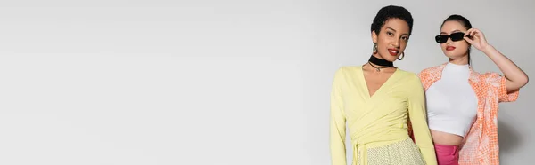 Copines souriantes multiethniques dans des vêtements élégants posant sur fond gris, bannière — Photo de stock