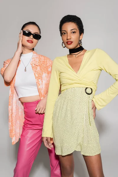 Mujeres jóvenes multiétnicas de moda posando en gafas de sol y ropa brillante sobre fondo gris - foto de stock