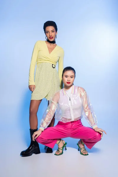 Positive interrassische Freundinnen in stilvollen Outfits posieren auf blauem Hintergrund — Stockfoto