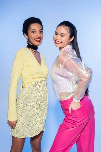 Fröhliche multiethnische Frauen posieren und blicken in die Kamera auf blauem Hintergrund — Stockfoto