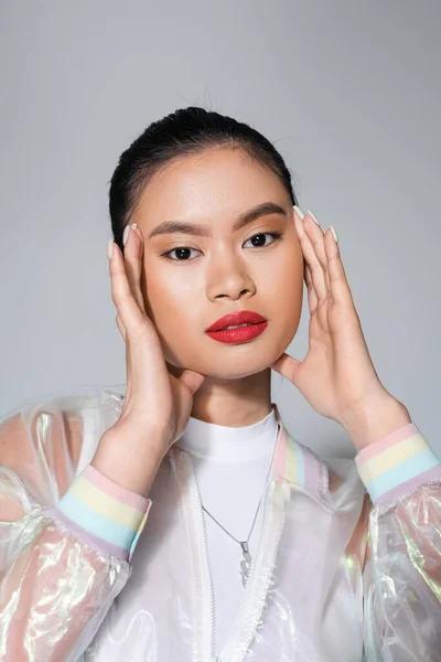 Retrato de joven modelo asiático con labios rojos tocando la cara aislada en gris - foto de stock