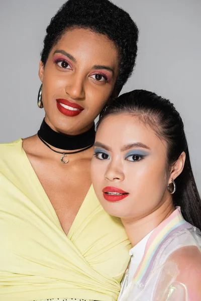 Позитивні багатоетнічні подружки з яскравим макіяжем дивиться на камеру ізольовані на сірому — стокове фото