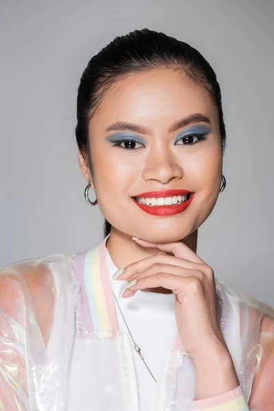 Lächelndes asiatisches Model mit buntem Make-up, das das Kinn berührt, isoliert auf grau — Stockfoto
