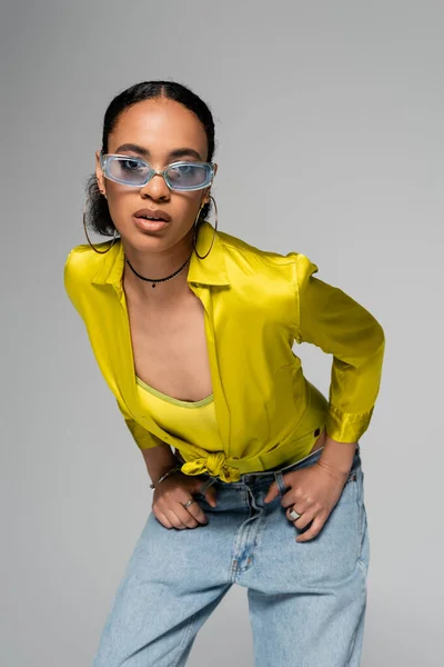 Jeune femme afro-américaine en lunettes de soleil bleu tendance posant isolé sur gris — Photo de stock