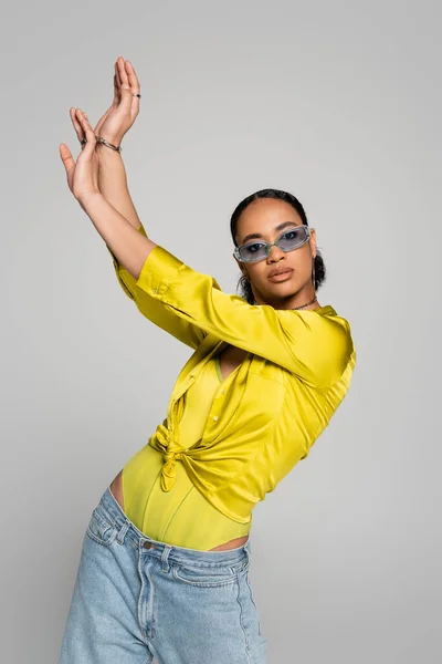 Pretty african american woman in blue sunglasses and stylish outfit posing with raised hands isolated on grey — Stock Photo