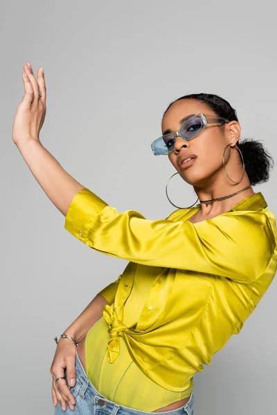 Encantadora mujer afroamericana en gafas de sol azules y elegante traje posando con la mano extendida aislada en gris - foto de stock