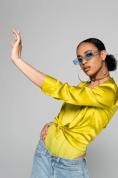 Pretty african american woman in blue sunglasses and stylish outfit posing with outstretched hand isolated on grey — Stock Photo